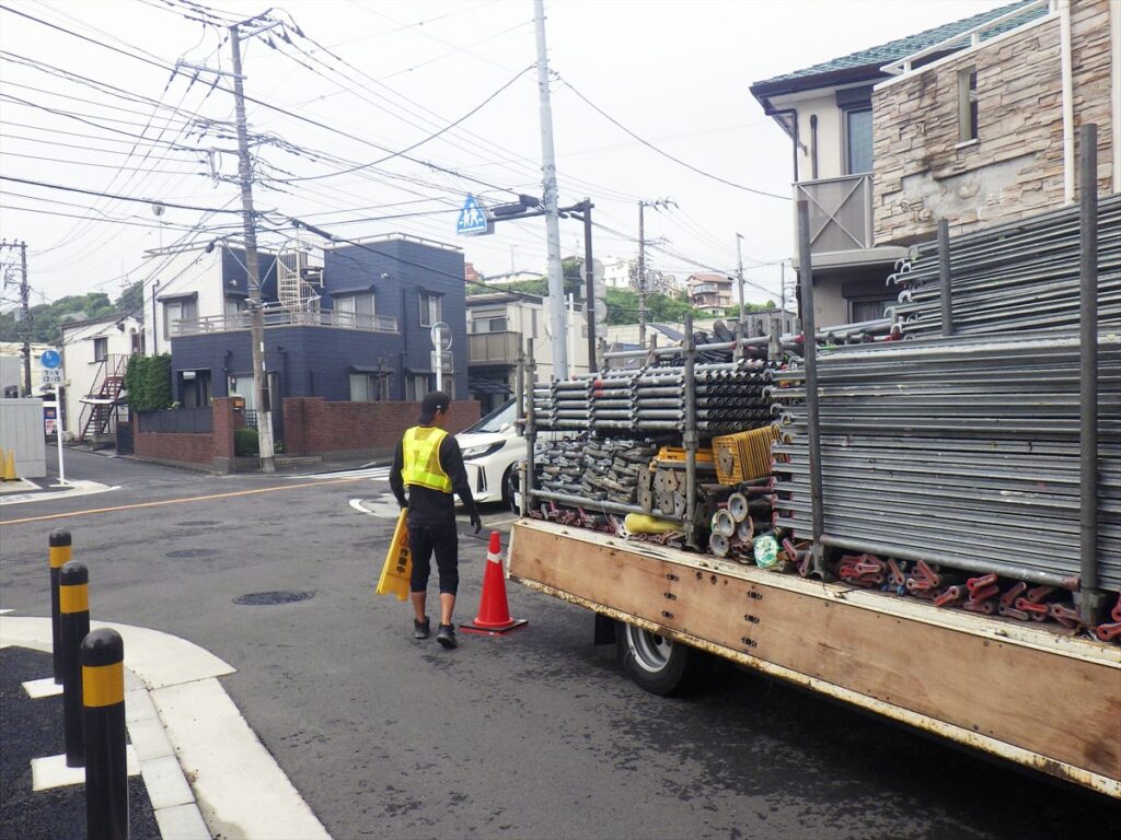 足場資材を積んだ足場屋のトラックと交通誘導員が立っている様子
